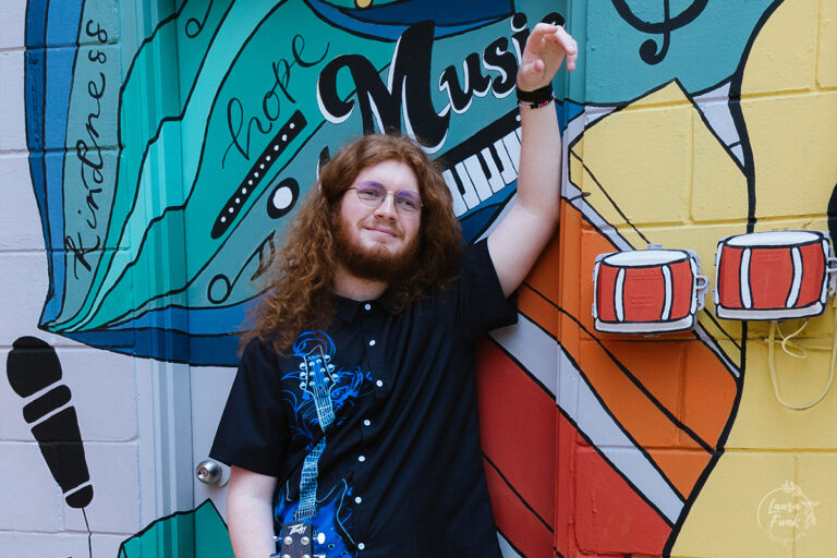 Guy leaning against a mural on a building posing with his guitar