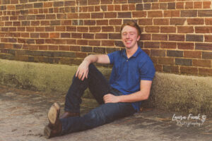 young man posing for senior portrait sitting on the sidewalk against a brick wall